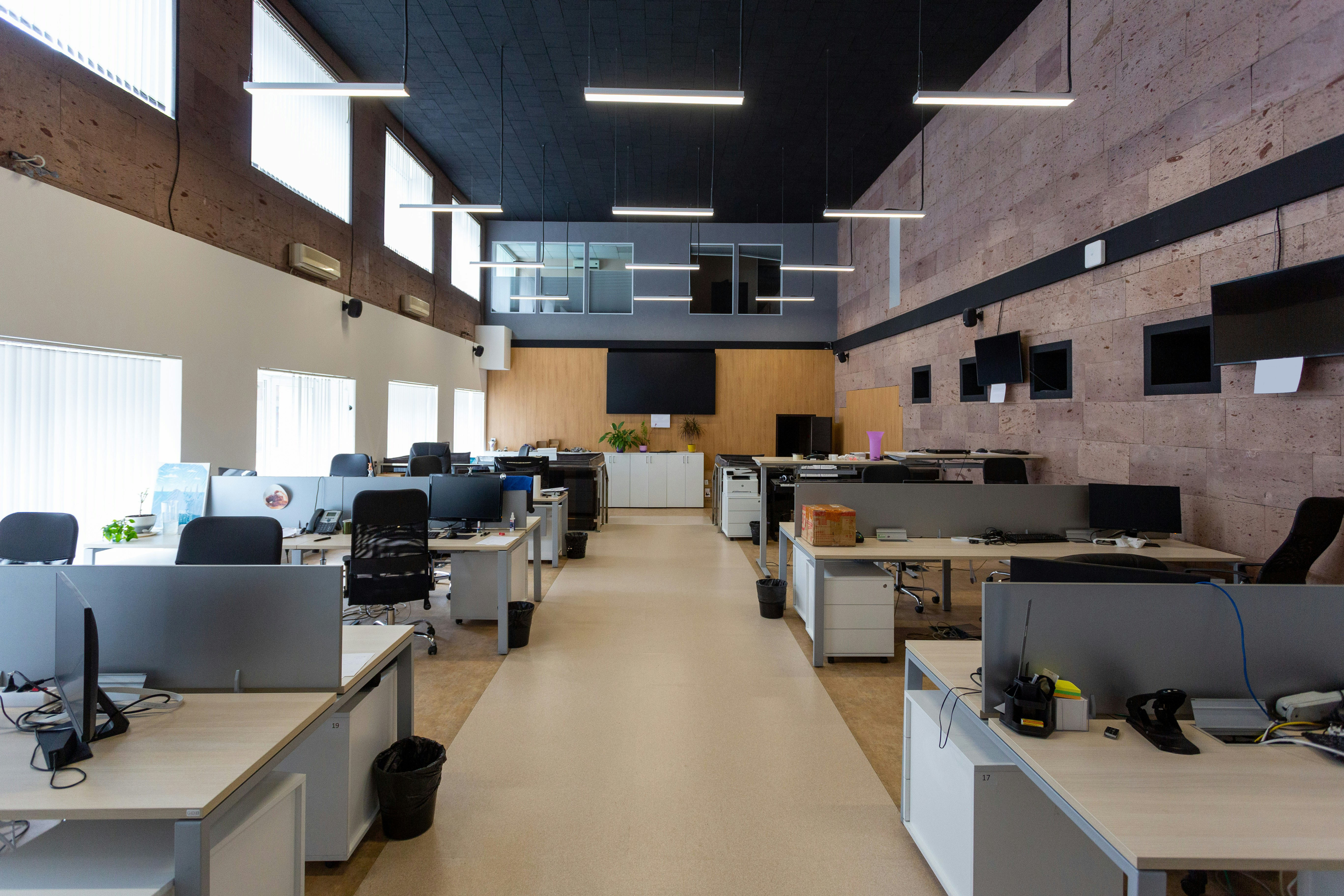 Modern commercial office build-out with rows of collaborative desks, suspended lights, textured accent wall, and ceiling-mounted monitors.